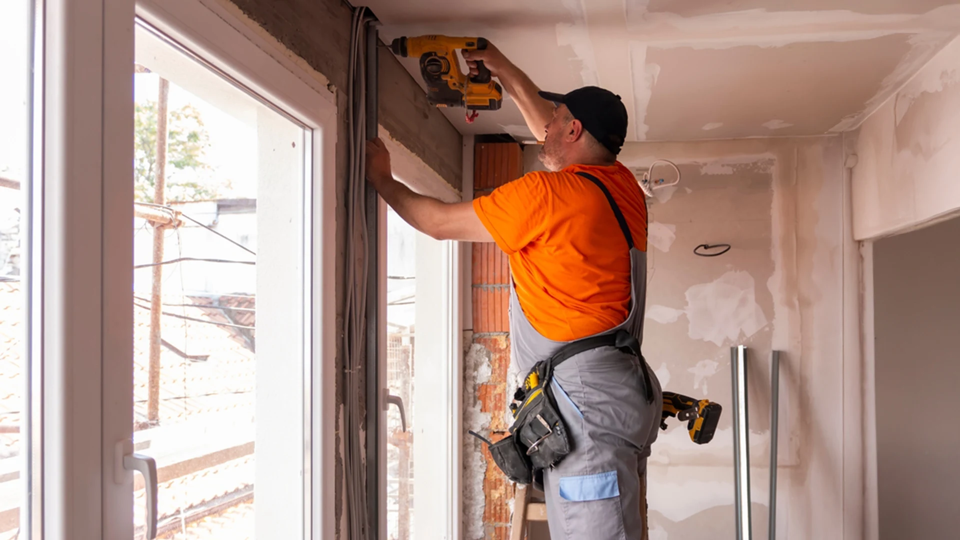 Tradesperson installing wall framing during a home renovation, using a power drill inside a partially finished room with exposed brick and plaster.