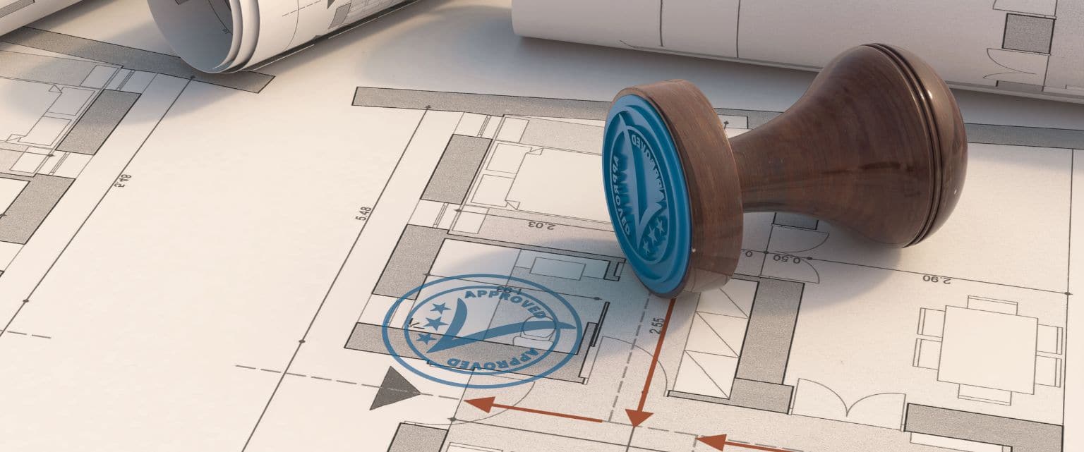 The image shows a wooden rubber stamp resting next to an approved construction blueprint, representing the building permit process.